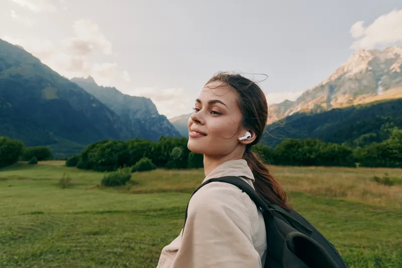 Young woman with long brown hair enjoys a peaceful moment outdoors in the mountains while wearing wireless earbuds and a light jacket with a backpack.
