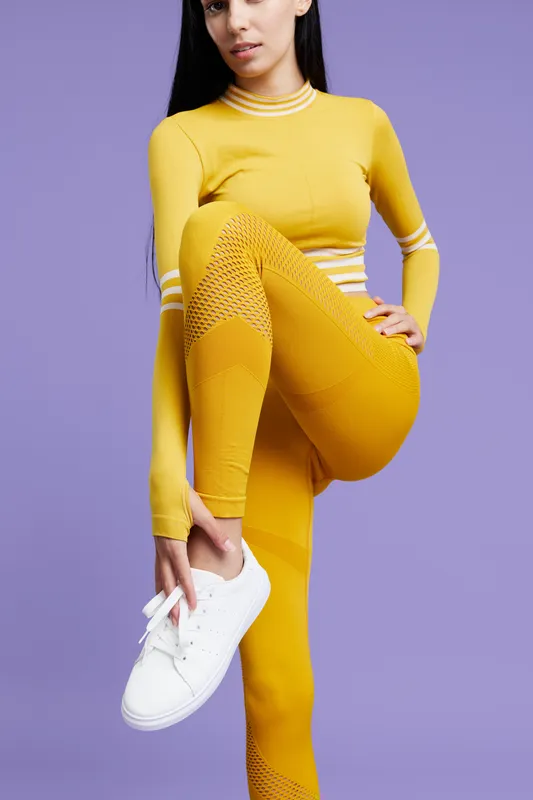 woman sport fitness in yellow activewear tying white sneakers on purple background, posing for workout, healthy lifestyle concept