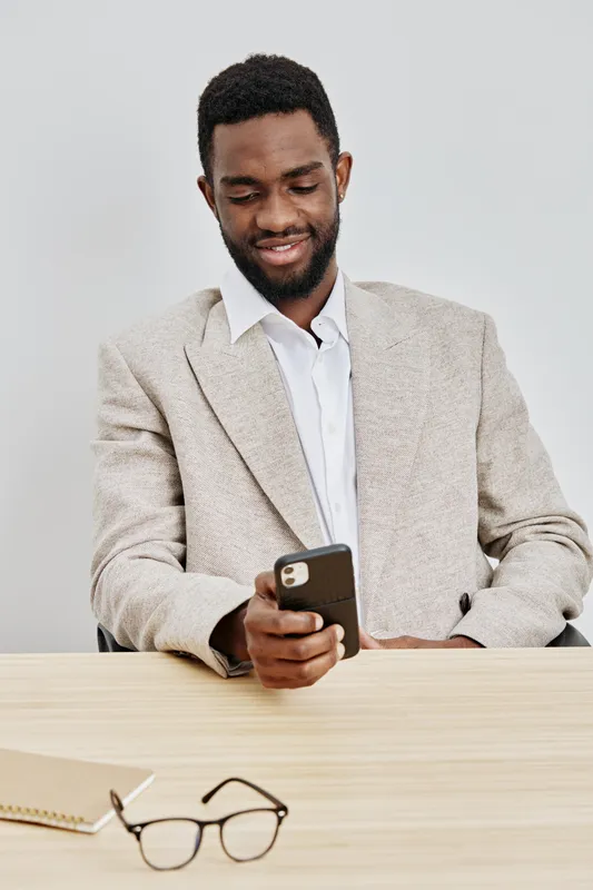 Smiling man using smartphone at desk, dressed in beige blazer and white shirt, in modern office setting, focused on screen, casual professional mood, indoor lifestyle concept, clean background, profes