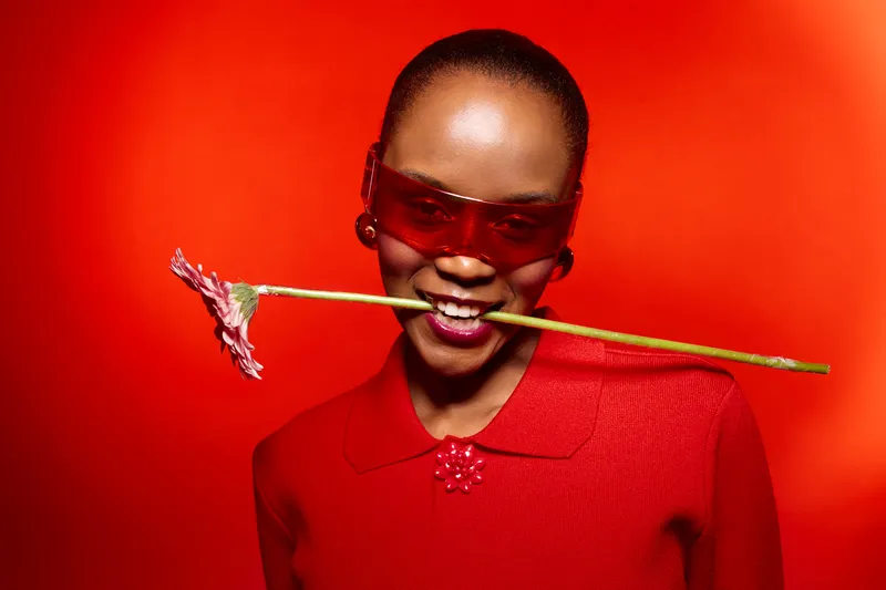 Smiling dark skinned African woman wearing red blouse and sunglasses holds flower stem in mouth on red background. Concept of fashion, joy, and vibrant color contrast with confident mood.