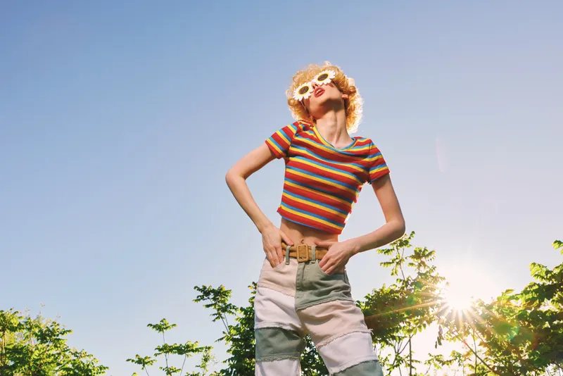 Sunny outdoor scene shows a woman wearing a striped rainbow crop top and jeans, posing confidently in bright daylight, conveying lifestyle, summer, sunshine, casual, retro color, and fashion.