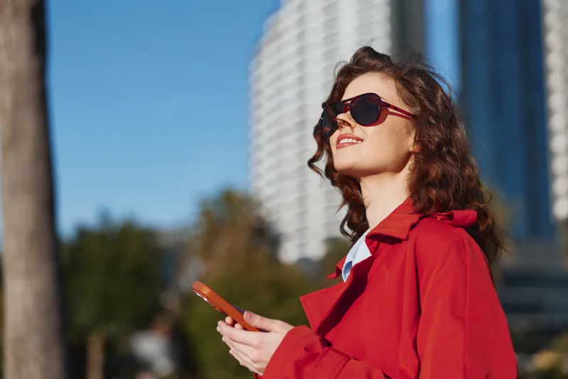 A confident woman in a red coat and sunglasses holds a smartphone outdoors with a blurred cityscape background on a sunny day.