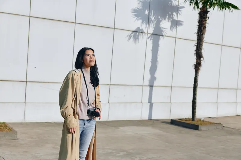 Woman with camera standing outdoors near a white wall, wearing a beige coat and denim, sunlight creates soft shadows, urban street photography mood and relaxed fashion vibe.