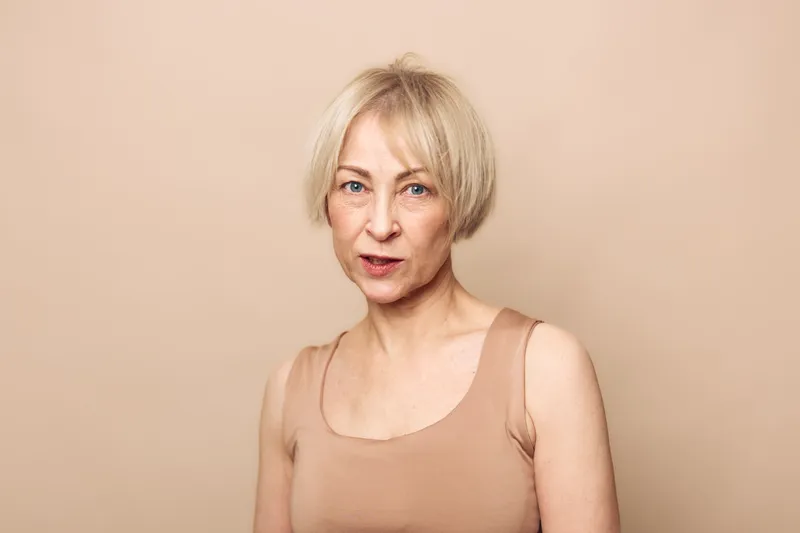 Portrait of a mature blond woman with short hair wearing a beige tank top against a soft beige background. Natural look, confident expression, minimalistic style, beauty and aging concept.