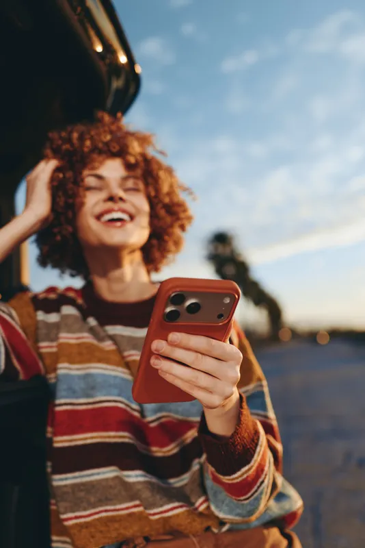 Woman lifestyle curly hair smiling smartphone outdoor beach sunset casualwear, striped sweater, holding phone in hand, laughing in warm sunlight, shallow depth background