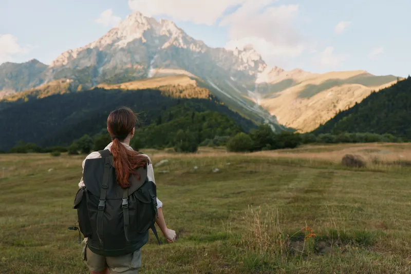 Woman hiker with backpack stands in a grassy valley, mountains ahead under a sunlit sky, conveying adventure and calm in a natural outdoor setting