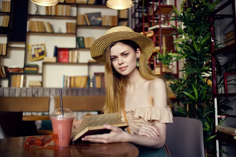 Симпатичная женщина в шляпе с книгой в руках читает во время отдыха в кафе