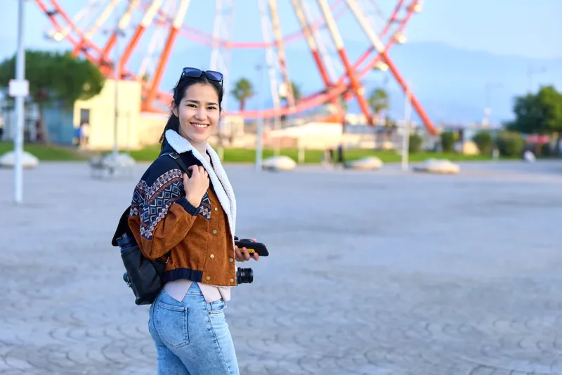 Woman with backpack at an outdoor amusement park on a sunny day, smiling beside a towering ride, wearing a casual jacket and jeans, ready for adventure and exploration