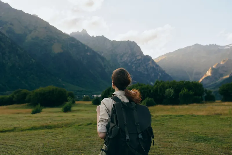 A woman with a backpack stands in a green field looking at distant mountains under a cloudy sky. Concept for lifestyle, travel, and outdoor adventure.