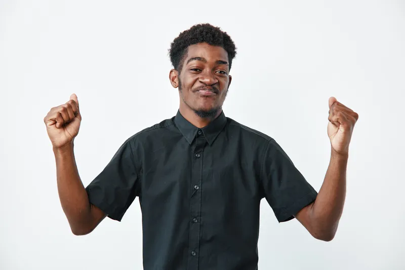 Young black man with dark skin wearing black shirt smiling and raising fists in celebration or success on white background. Positive emotion and confident expression.