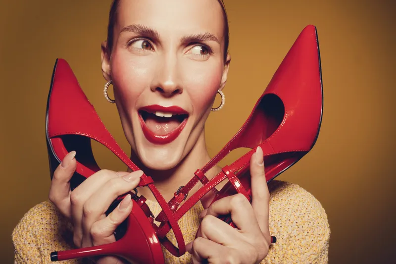 Excited woman holds red high heels near face with open mouth expression on yellow background. Concept of fashion, style, footwear, and joyful emotions in studio portrait.