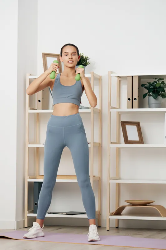 Fitness woman in blue athletic outfit lifts dumbbells in a bright home gym, performing a standing shoulder press with a focused expression, shelves and decor in the background.