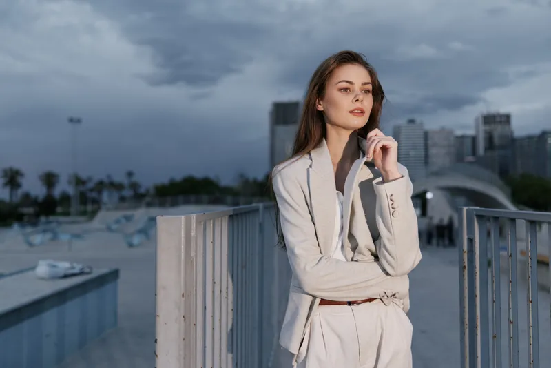 Confident woman in business attire posing near modern architecture, showcasing elegance and determination against a cloudy skyline.