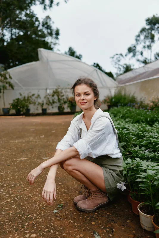 Young woman in casual outfit squatting in a botanical garden, surrounded by green plants, showcasing a bright and natural atmosphere perfect for wellness concepts