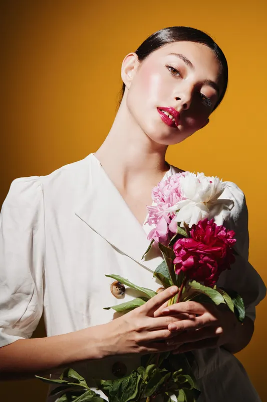 Young woman in elegant white outfit holds a bouquet of pink and white flowers against a rich yellow background. Soft lighting highlights fresh skin and glossy red lips. Concept of beauty fashion and