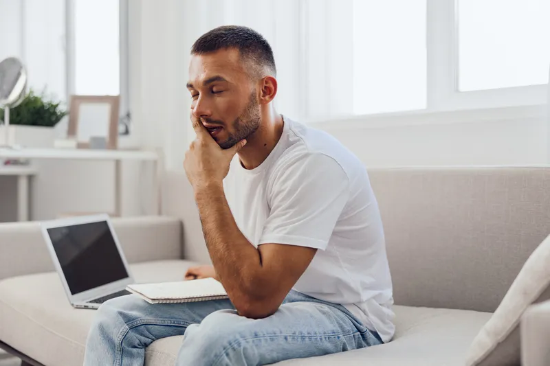 Thoughtful man sitting on the couch with a laptop, reflecting on ideas while holding a notebook, showcasing productivity and inspiration in a modern living space.