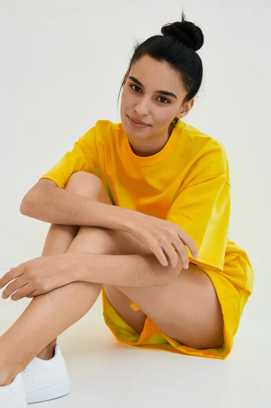 young woman sitting relaxed in bright yellow casual clothing with white sneakers on plain light background, smiling and looking at camera confidently