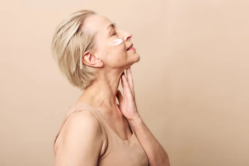 Mature woman with short blonde hair applies white under eye cream, gently touching neck and smiling with closed eyes. Concept of skincare, beauty routine, anti aging, self care, and wellness.