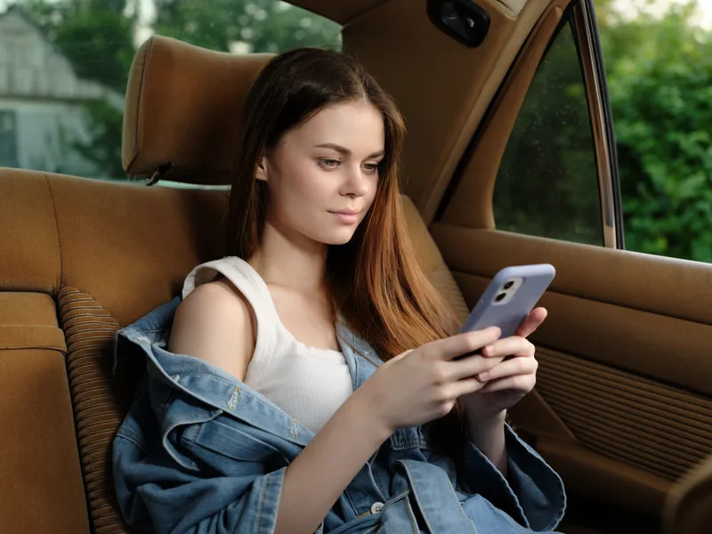 Young woman with long hair in car using smartphone, wearing casual denim jacket and tank top, serene expression, green background, modern lifestyle, technology integration.