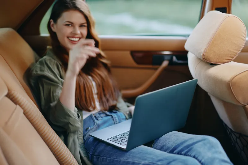 Young woman in casual attire, smiling and working on a laptop in a vintage car, showcasing relaxation and productivity in a natural setting.