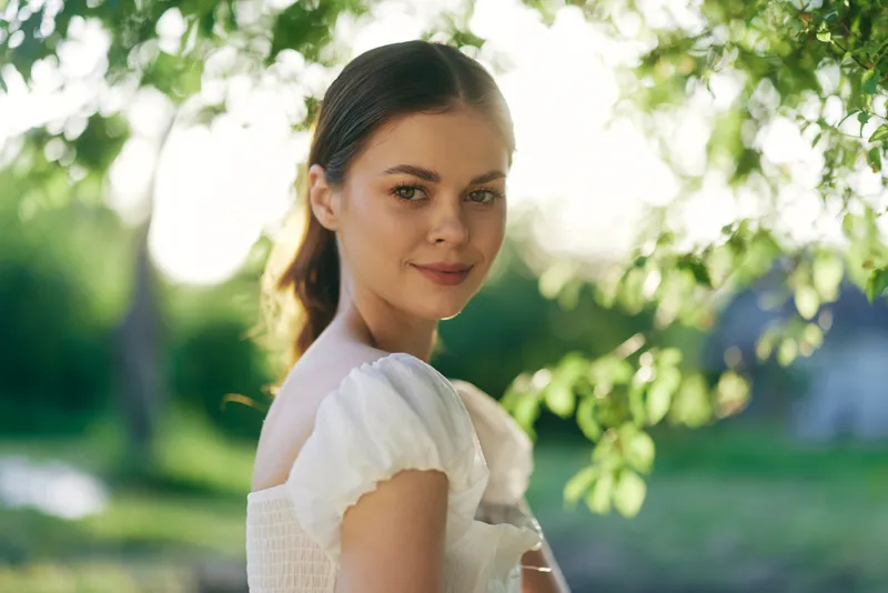 Beautiful young woman in white dress smiling in sunny outdoor setting, surrounded by green leaves, evoking a sense of joy and nature
