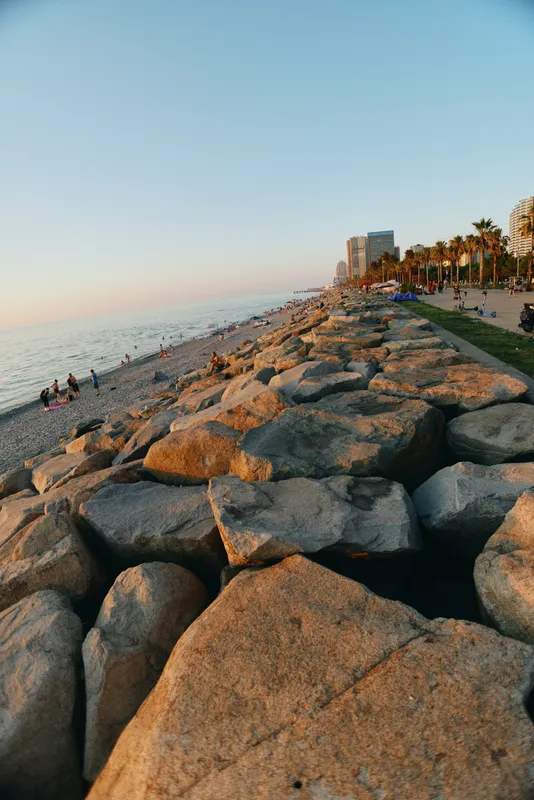 rocky beach coastline with calm sea and sunset lighting along promenade walkway and palm trees under clear sky in warm evening atmosphere