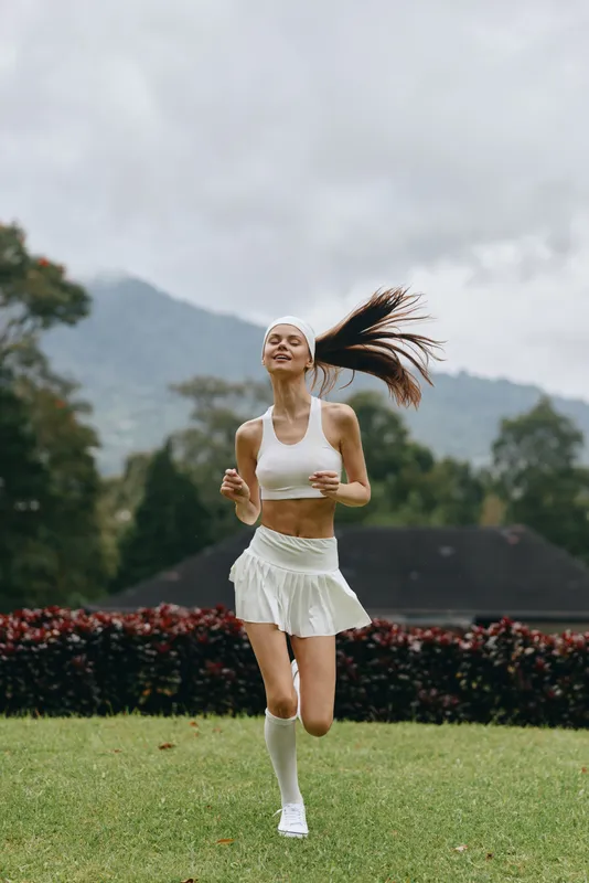 Young athletic woman jogging outdoors in white sportswear, showcasing fitness and vitality, amidst a lush green backdrop, perfect for active lifestyle concepts