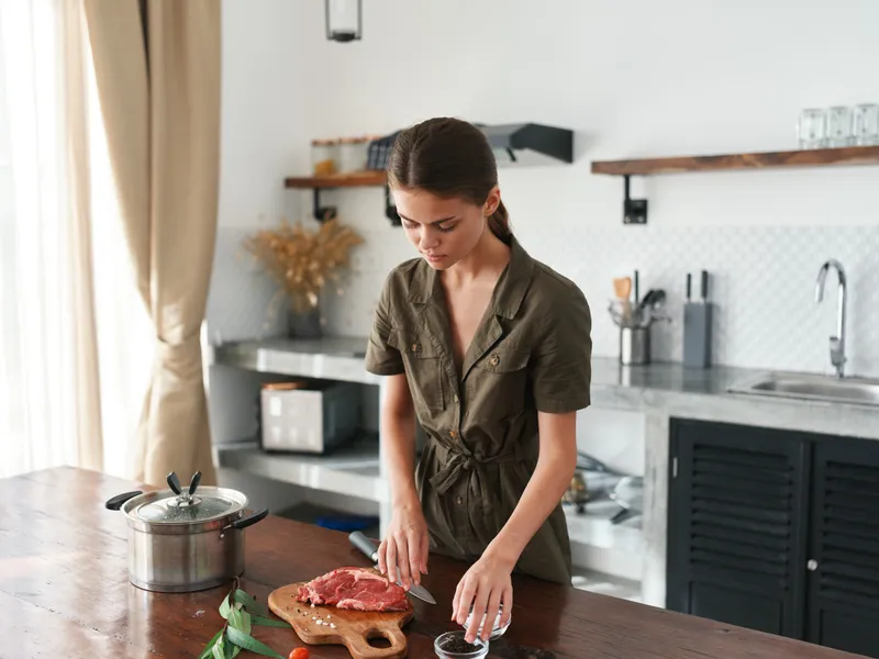 Женщина на кухне готовит обед из мяса, овощей и специй на деревянном столе на фоне стильной кухни