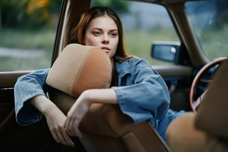 Young woman in denim jacket leaning in a vintage car, expressing serene emotions with a soft background and warm, natural colors, perfect for lifestyle themes.