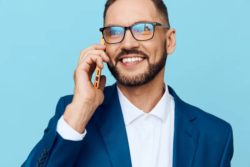 A professional man in a blue suit stands against a solid blue background, engaged in a phone call, showing confidence, focus, and a positive mindset in a business setting.