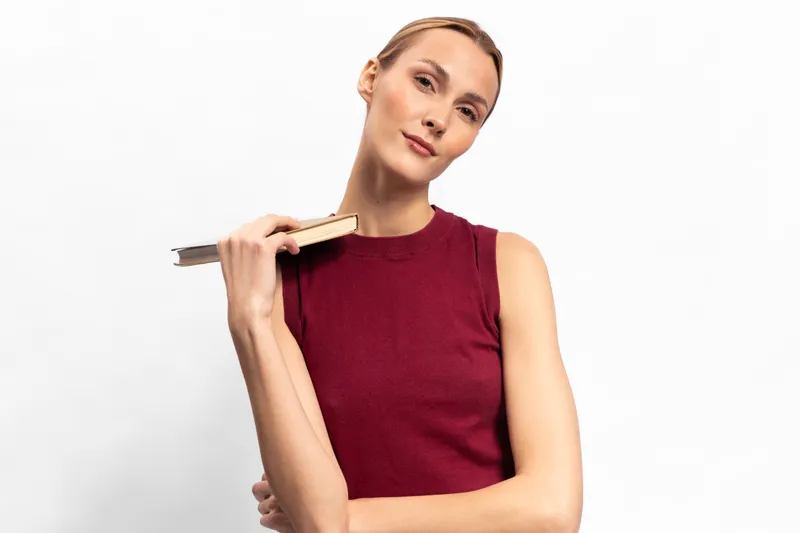 Confident young woman in a burgundy sleeveless top holds a book on her shoulder, posing against a white background. Concept for education, knowledge, and casual style.