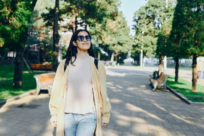 Street fashion of a woman in a light coat and jeans walking through a sunlit park, sunglasses on, relaxed stride, urban nature setting, fashionable outdoor moment ideal for lifestyle and travel