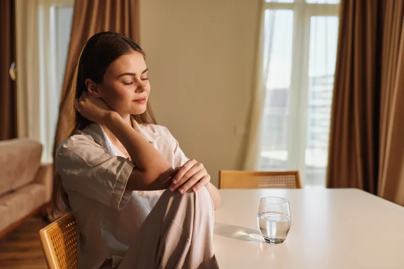 Woman portrait relaxation home glass table - Young woman sits at table with a glass of water, eyes closed and hand on neck in warm sunlight, calm morning and quiet wellbeing routine.