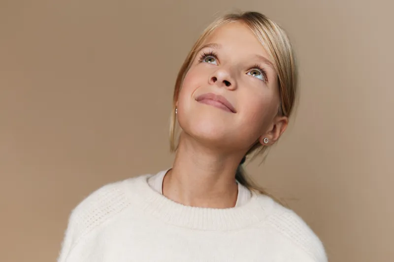 Close-up portrait of a thoughtful woman gazing upward against a warm beige backdrop, wearing a soft knit sweater. Calm, hopeful mood with gentle lighting and relaxed expression.