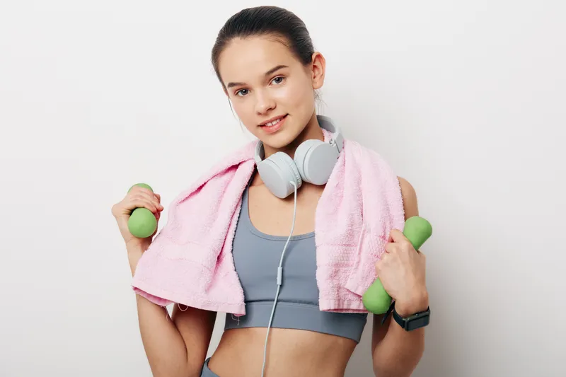 Fitness, woman, workout, dumbbells in hands with green weights, gym ready, wearing gray sports top and pink towel, headphones around neck, focused expression after training.