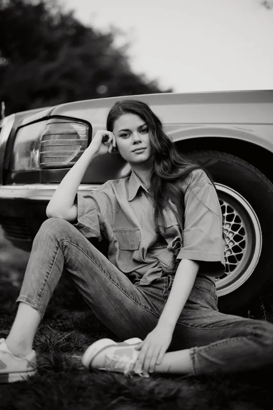 Black and white portrait of a young woman in oversized shirt and jeans, sitting beside a vintage car, showcasing a casual and relaxed style.