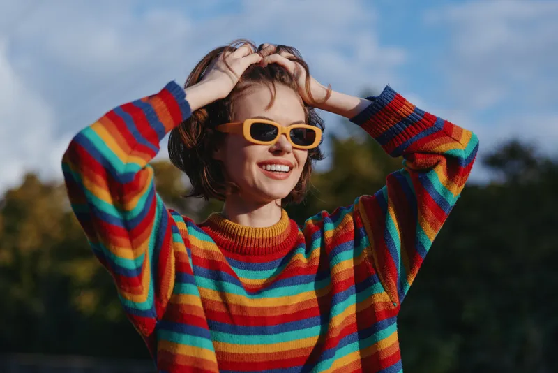 Young woman in a colorful striped sweater and yellow sunglasses smiling outdoors, hands in hair, casual outfit in park setting, joyful lifestyle portrait for fashion or travel.