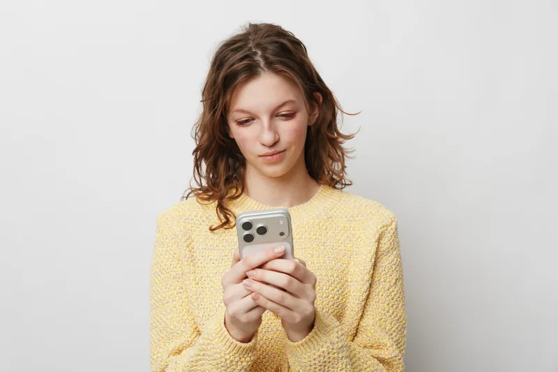 Young woman in yellow sweater looks at smartphone with slight smile, standing against plain light background. Concept of communication, technology, social media, and casual lifestyle.