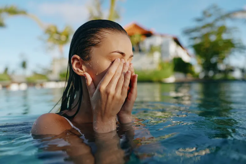 A young woman with wet hair relaxes in an outdoor pool under natural sunlight, gently touching her face. The background shows blurred greenery and a house, lifestyle concept.