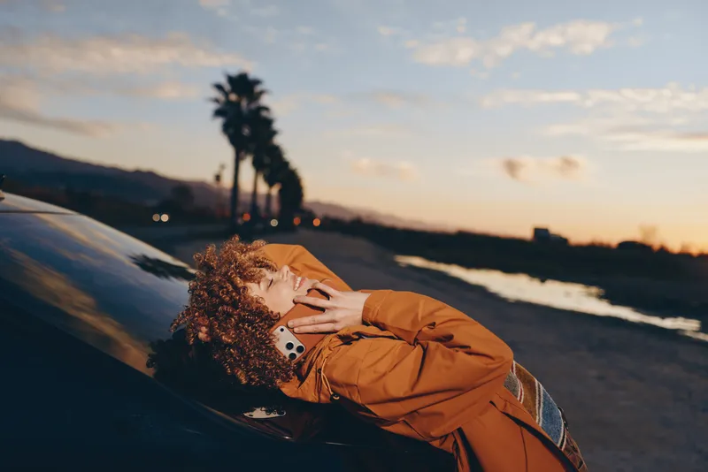 Woman lifestyle outdoors sunset portrait orange jacket smiling with eyes closed, hands on hair, coastal hillside trail, warm evening light, soft breeze, natural colors, cheerful mood