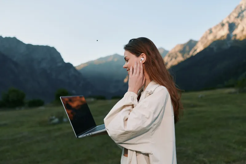 A woman stands outdoors in a scenic valley, holding a laptop near her ear as she talks, wearing a light coat. The scene conveys focus, openness, and modern remote work in nature.