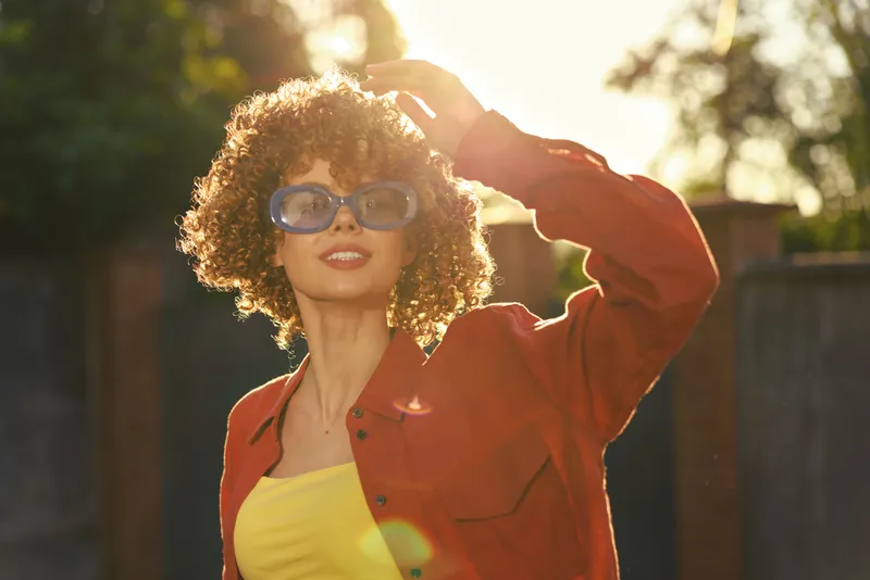 A stylish woman outside on a bright sunny day wearing a yellow top and red jacket, posing with oversized sunglasses, capturing confident, carefree lifestyle moments in warm golden light