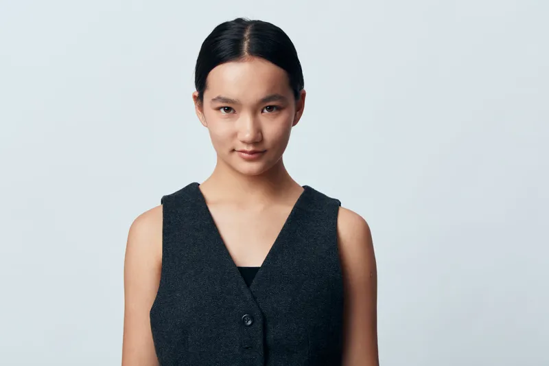 Portrait of confident woman with dark hair wearing sleeveless dark vest looking at camera against plain background, studio lighting emphasizing facial features and expression.