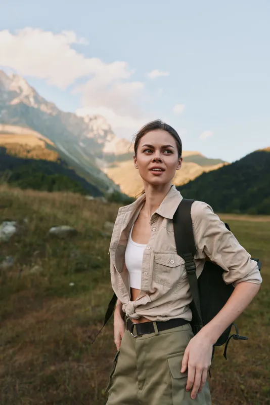 Cheerful woman outdoors, hiking lifestyle with a backpack, standing in a meadow and looking ahead, sunlit moment, casual attire, natural colors, mountain backdrop, lush greenery and open sky