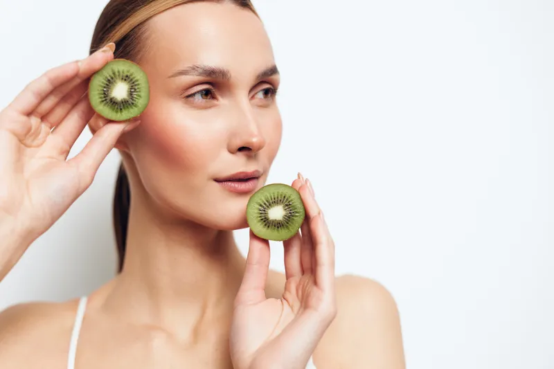 A young woman holds two halves of a kiwi fruit near her face, looking away thoughtfully against a white background. Concept for healthy lifestyle and natural beauty.