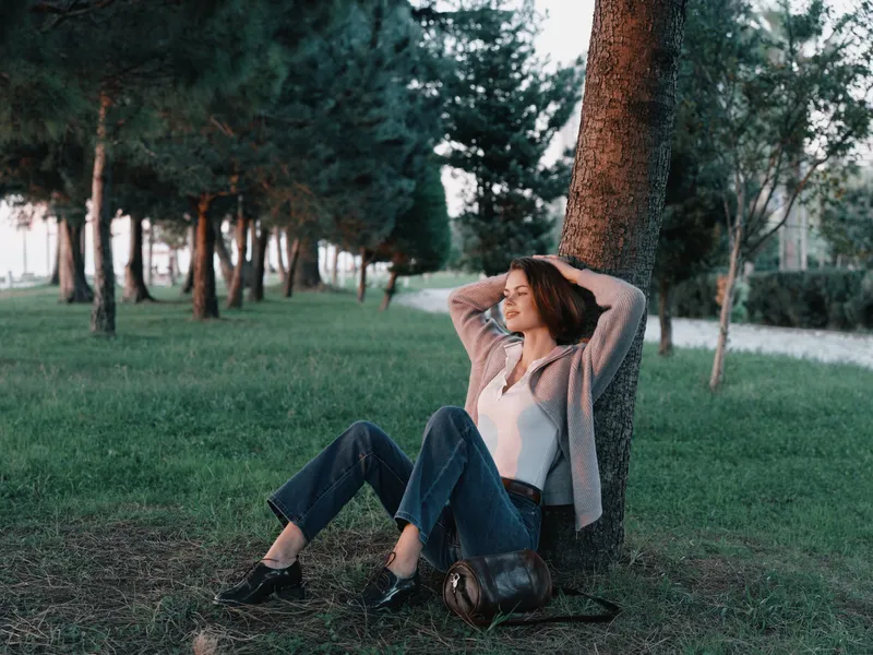 A relaxed woman rests beside a tree in a park, smiling softly as she gazes ahead, creating a tranquil and natural atmosphere that feels calm, peaceful, and inviting.