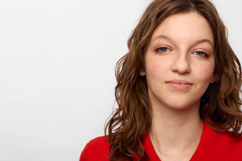 Young woman with natural makeup and freckles looks confidently at camera. Casual hairstyle and red top create simple, fresh portrait on white background. Concept of beauty and self-confidence.