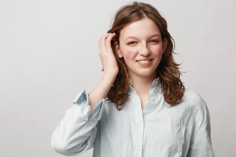 Smiling young woman with curly brown hair wearing a light blue shirt touches her hair. Studio portrait on white background. Concept of natural beauty, casual style, and positive mood.