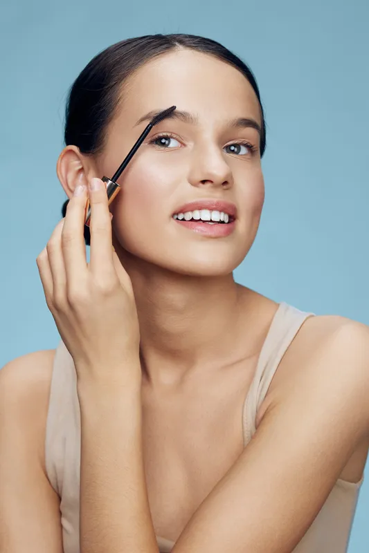Young woman applying eyebrow gel with a cheerful smile, showcasing beauty and cosmetics against a light blue background in a minimalistic style