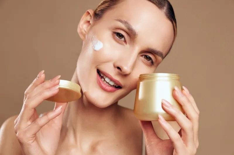 Young woman with brunette hair applies cream from a gold jar in a close up portrait, soft lighting, smooth skin, calm expression, warm beige studio background during skincare